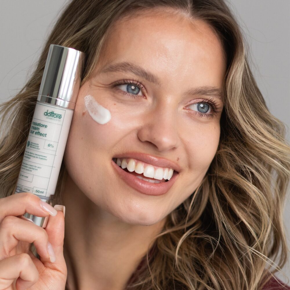 Uśmiechnięta modelka trzyma Rossatore Cream Blur Effect, kropla kremu na policzku