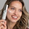Uśmiechnięta modelka trzyma Rossatore Cream Blur Effect, kropla kremu na policzku