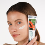 Młoda dziewczyna prezentująca konsystencję peelingu na policzku i opakowanie produktu przy twarzy