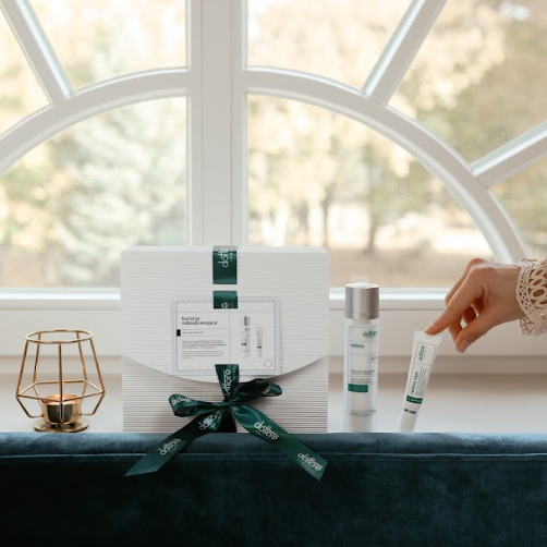 Dłoń sięgająca po tubkę kremu pod oczy z zestawu prezentowego z retinolem stojącego na oknie