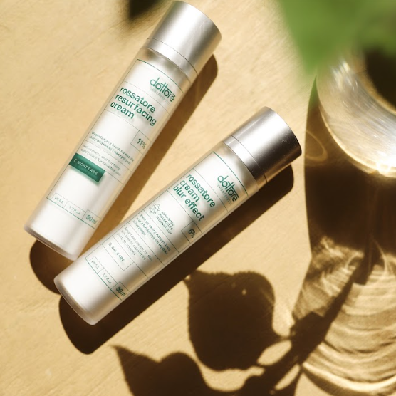 Dwie butelki kremów Rossatore do cery naczynkowej leżące na beżowym tle w cieniu roślin