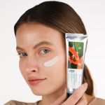 Młoda dziewczyna prezentująca konsystencję peelingu na policzku i opakowanie produktu przy twarzy