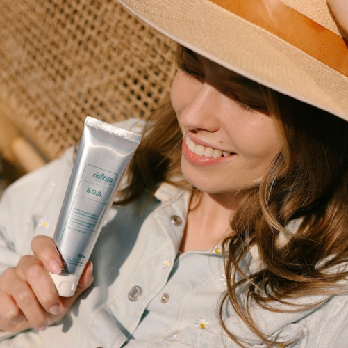 Kobieta w słomkowym kapeluszu trzymająca łagodzący krem SOS, chroniąc twarz przed słońcem.