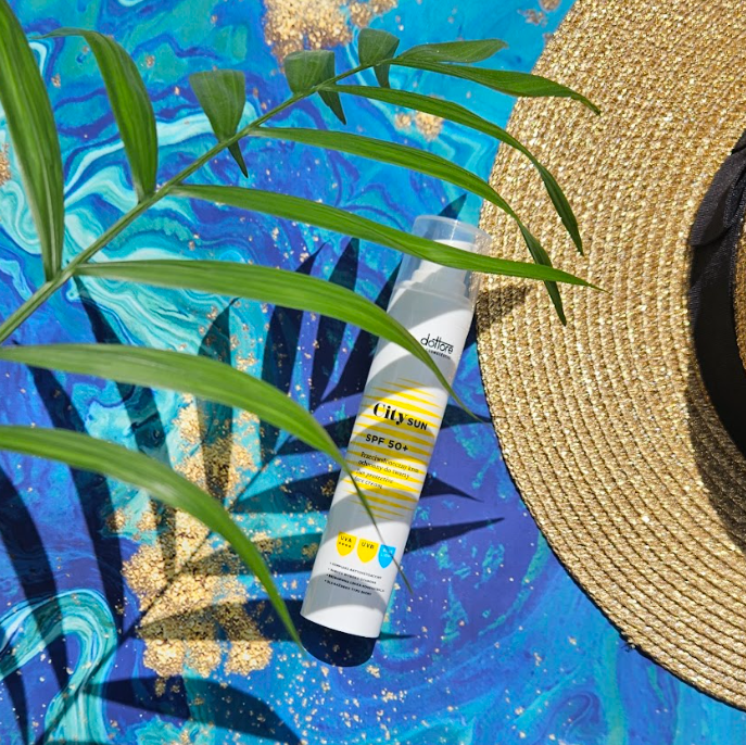 Krem ochronny SPF 50+ leżący na niebieskim tle w liście palmy obok słomkowego kapelusza.